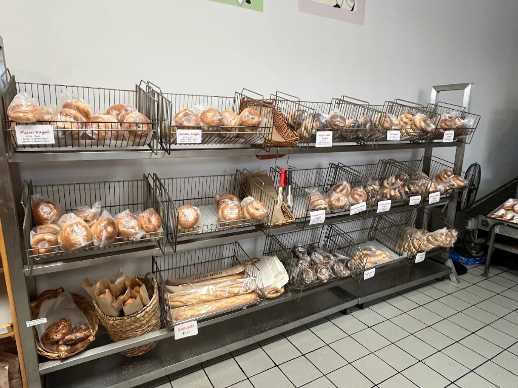 Metal shelving stacked with bagels in baskets: plain, sesame, egg, and other varieties with price cards.