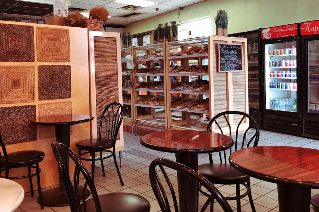 Dining area with wood tables, black chairs, wicker wall panels, and beverage cooler with Happy Bagel Bakery logo.
